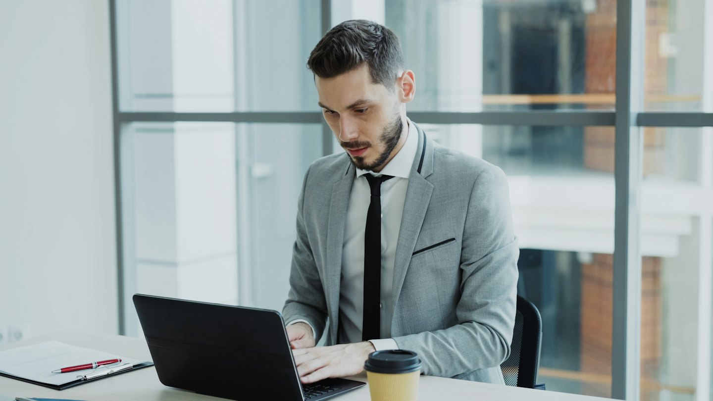 Man in a grey suit works on a laptop at a desk.